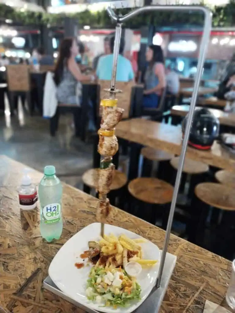 Leckerer Fleisch- und Gemüsespieß in einem Restaurant. Der Spieß hängt an einem Ständer und wird mit Salat und Pommes auf einem Teller serviert. Das Restaurant hat eine lebendige, moderne Atmosphäre.