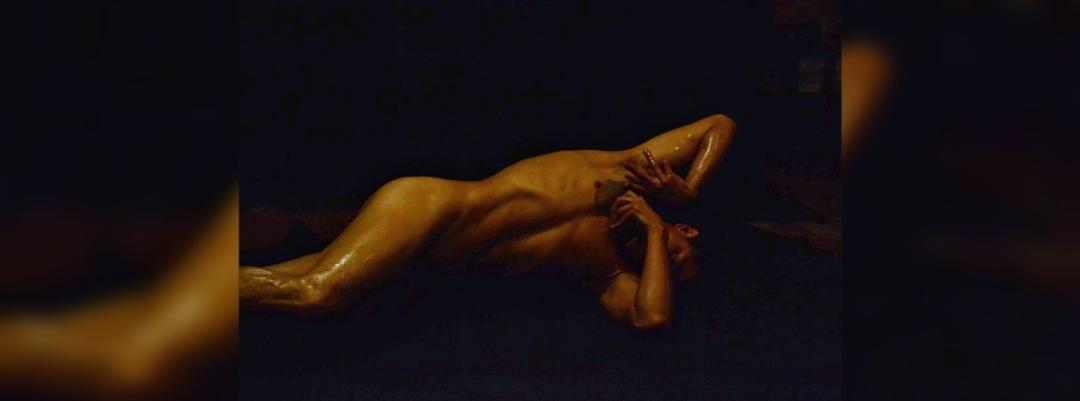 Hombre desnudo en posición artística sobre fondo oscuro, con iluminación suave resaltando su figura y musculatura, en una escena que invita a la reflexión y la expresión artística.