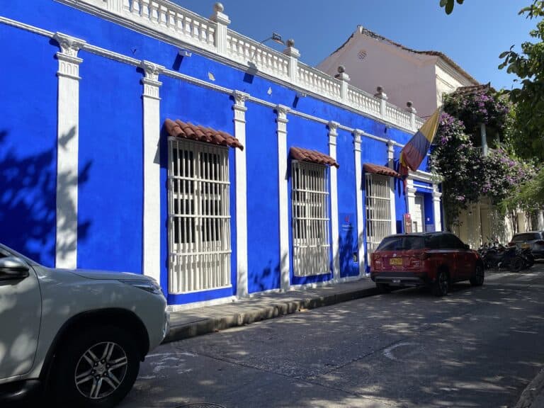 Colorida fachada azul y blanca de un edificio histórico en un día soleado en Bogotá, con vehículos estacionados y árboles con flores, ideal para eventos culturales y turísticos.
