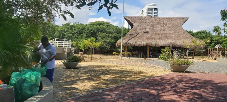 Basurero en parque infantil con estructura de palapa y áreas verdes, promoviendo limpieza y conservación del espacio para eventos y actividades al aire libre en Colombia.