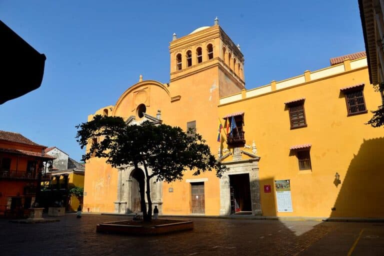 Arco y fachada de iglesia colonial en San Juan, Colombia, con cielo azul y árbol en primer plano, perfecta para eventos culturales y turísticos.