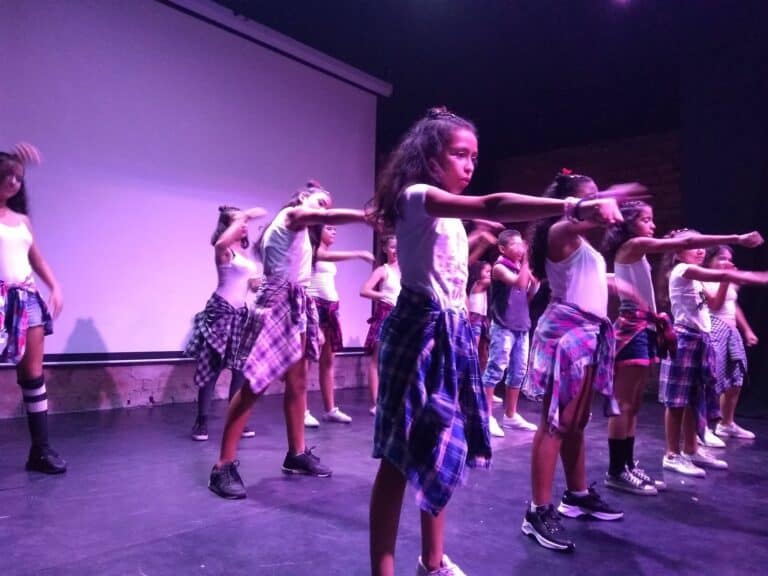 Bailarinas infantiles en presentación de danza con vestimenta casual y pañuelos a cuadros en un escenario con fondo de pared morada y luces de escenario, mostrando talento y coordinación.