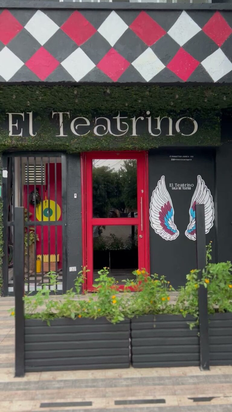 Puerta de entrada de El Teatrino, teatro y sala de eventos culturales, con decoración moderna en negro y rojo, y alas de ángel pintadas en la pared, rodeada de vegetación y flores.