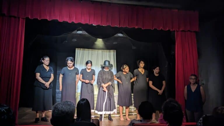 Personas actuando obra de teatro en escenario con cortinas rojas.