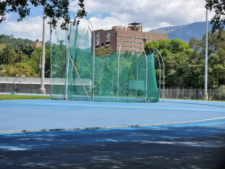 Upper text: Outdoor running track with safety enclosure and training area, located in a natural setting with trees, mountains, and a building in the background.
