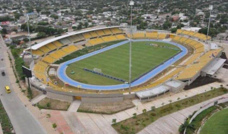 Ingrese la captura del estadio en vista aérea en color, con asientos amarillos, césped y pista de atletismo, en una zona urbana.