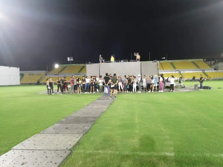 A group of people gathered in a stadium at night, apparently participating in an event or concert production, with a stage and lights in the background.