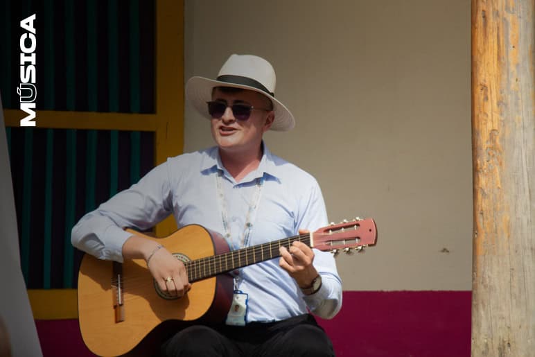 Guitarrista tocando en un evento musical al aire libre, vestido con sombrero y gafas de sol, disfrutando de la música en un ambiente festivo y vibrante.