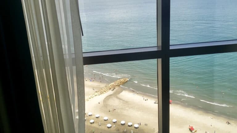 Hermosa vista de la playa desde una habitación con ventana de cristal, mostrando arena, olas y personas disfrutando del mar y sombrillas en un día soleado.