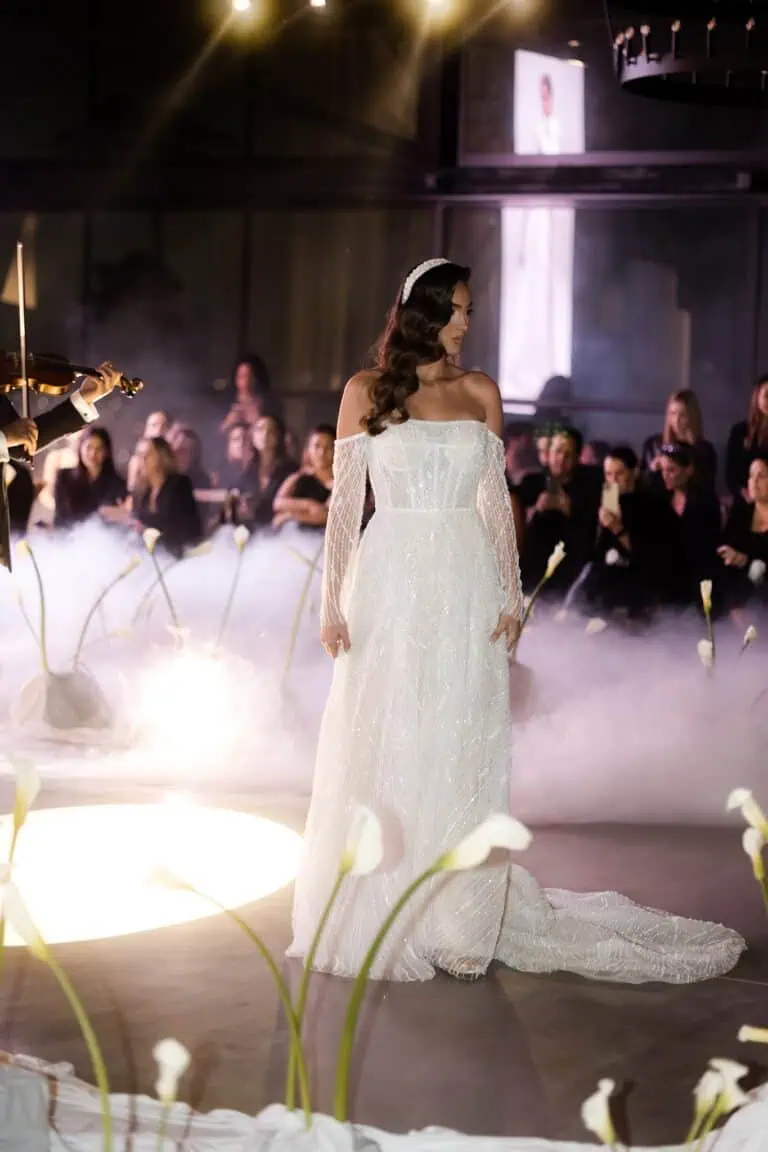 一件优雅的长款婚纱出现在婚纱时装秀的T台上。一位模特身穿白色蕾丝礼服，礼服上点缀着闪亮的细节，周围环绕着助手，现场还有音乐表演，整个时装秀的场地布置得十分精致。