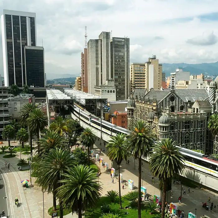 Catedral de Bogotá en el centro de la ciudad con tren en movimiento, rodeada de palmeras y edificios modernos, bajo un cielo parcialmente nublado.