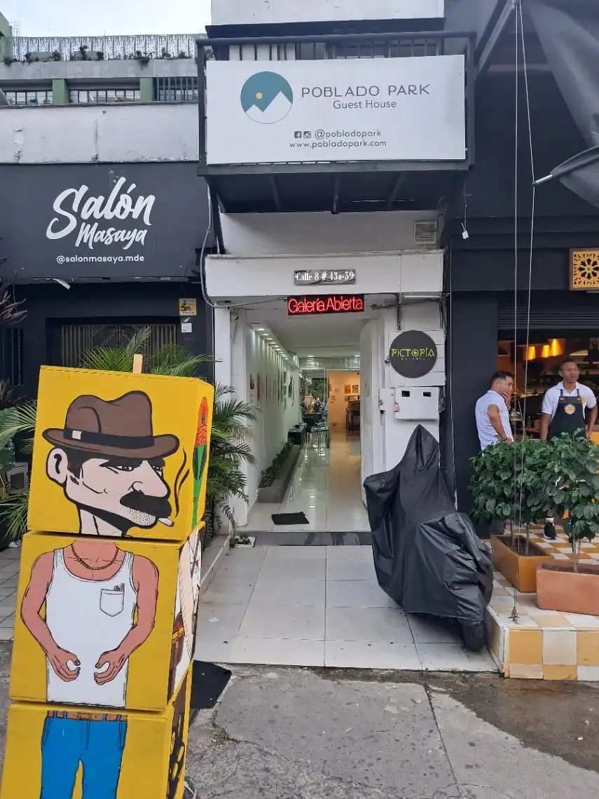 Imagen de un local en una calle, con cartel de Poblado Park Guest House y entrada a galería de arte, ambiente urbano y moderno, con decoraciones y personas en el exterior, ideal para eventos y turismo en Medellín.