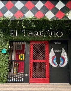 Entrada de teatro con muro decorativo y puerta de rejas rojas, cartel con alas y vegetación, fachada moderna y llamativa para eventos culturales y teatrales en Cartagena.