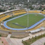 Estadio de fútbol con graderías amarillo y césped en perfecto estado, vista aérea del estadio en una ciudad con áreas residenciales alrededor.