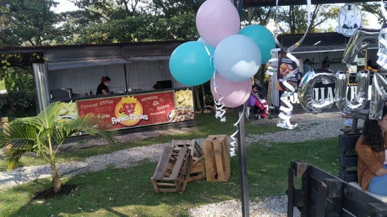 Arco de globos en tonos pastel con decoración para evento infantil en espacio al aire libre, con área de comida y ambiente festivo.