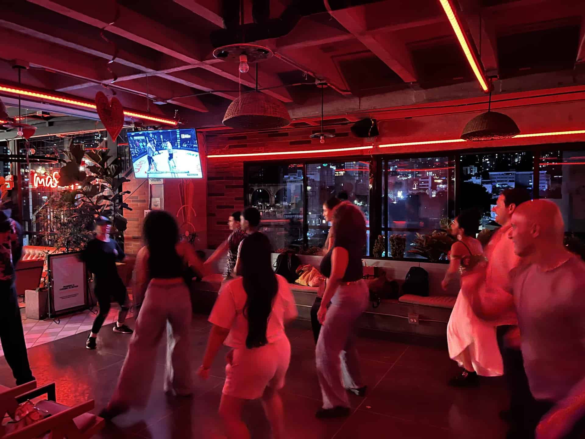 Diversión en evento con personas bailando en un ambiente vibrante y moderno, iluminado con luces rojas, con vista a la ciudad y transmisión en vivo en la pantalla.
