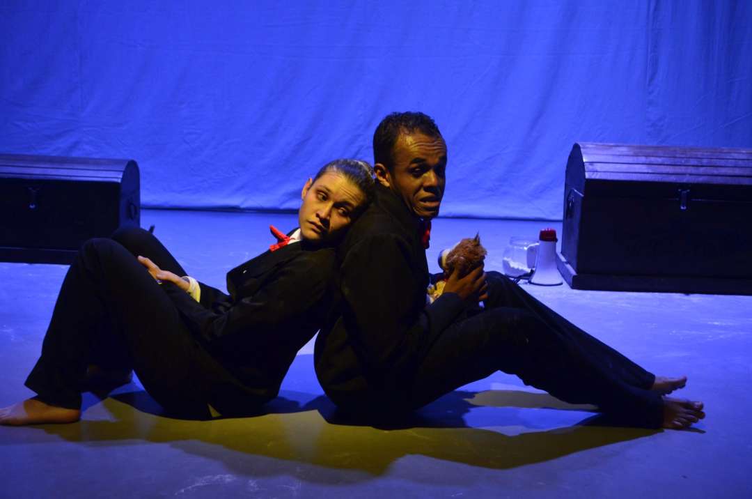 Seres humanos en una puesta en escena teatral con iluminación azul, actores en posición recostada en el escenario, expresiones emotivas, vestimenta negra y accesorios en un entorno de teatro profesional.