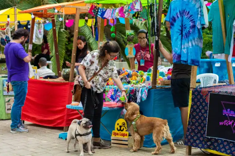 Artesanía y productos para mascotas en feria al aire libre en Colombia, vendedores con puesto de coloridos artículos, usuarios con perros, ambiente festivo y natural, evento de mercado artesanal y mascotas en celebración urbana.