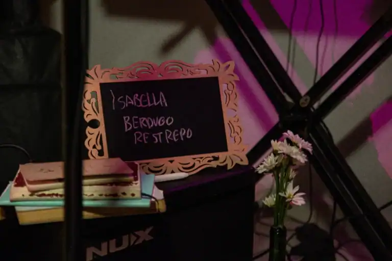 Bocadillo con lista de nombres en cartel decorativo y flores en vaso, en un evento social o celebración.