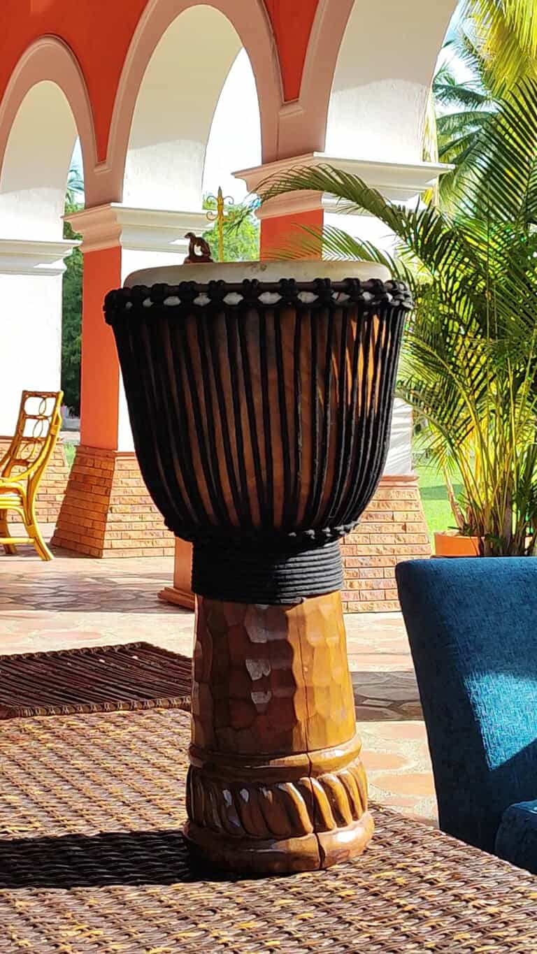 Batedor de madera tradicional en un espacio con arquitectura colonial y vegetación tropical en el fondo.