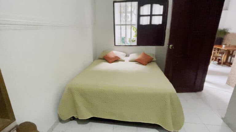 Dormitorio con cama doble, ropa de cama en color beige y almohadas rosas, ventana con rejas y vista a un patio, puerta de madera oscura y ambiente acogedor, en un espacio interior de alojamiento en Colombia.