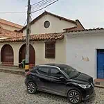 Una vecchia casa in un quartiere tradizionale con facciate bianche, tetto di tegole rosse, porta blu e finestre in argilla, su una strada acciottolata con un'auto nera parcheggiata e un palo della luce.