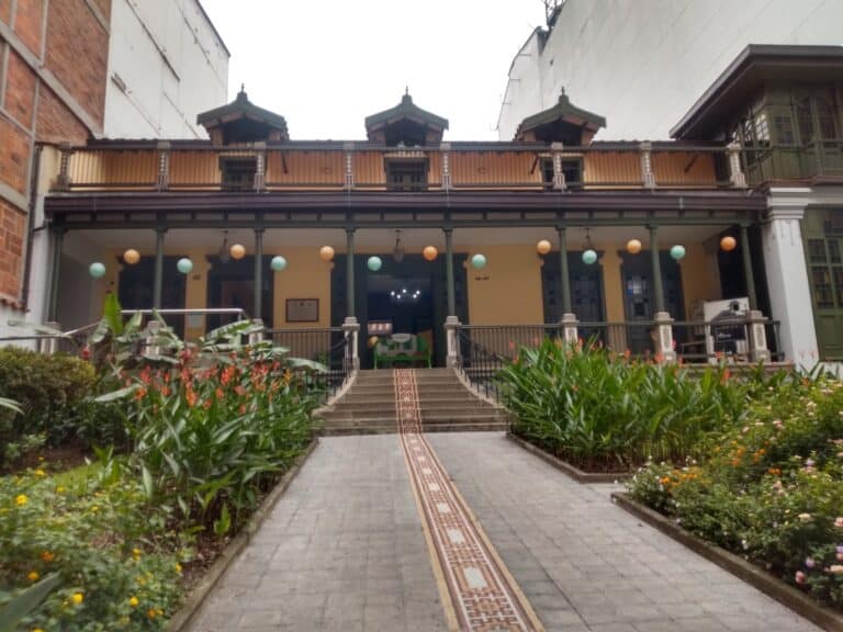 Detrás de una entrada elegante, un edificio histórico con balcones decorativos y una planta ornamental en el frontispicio, en un entorno urbano con jardines floridos y un sendero de entrada.