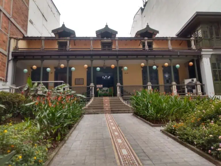 优雅的入口之后，是一座历史悠久的建筑，带有装饰性的阳台和门前的观赏植物，周围环绕着花园和入口小径，位于城市环境中。