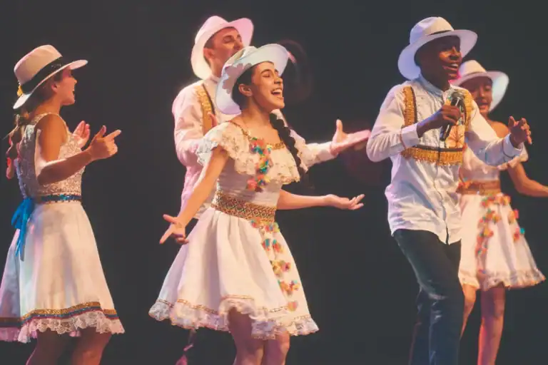 Bailarines y cantante en presentación musical con trajes folklóricos colombianos y sombreros blancos en escena.
