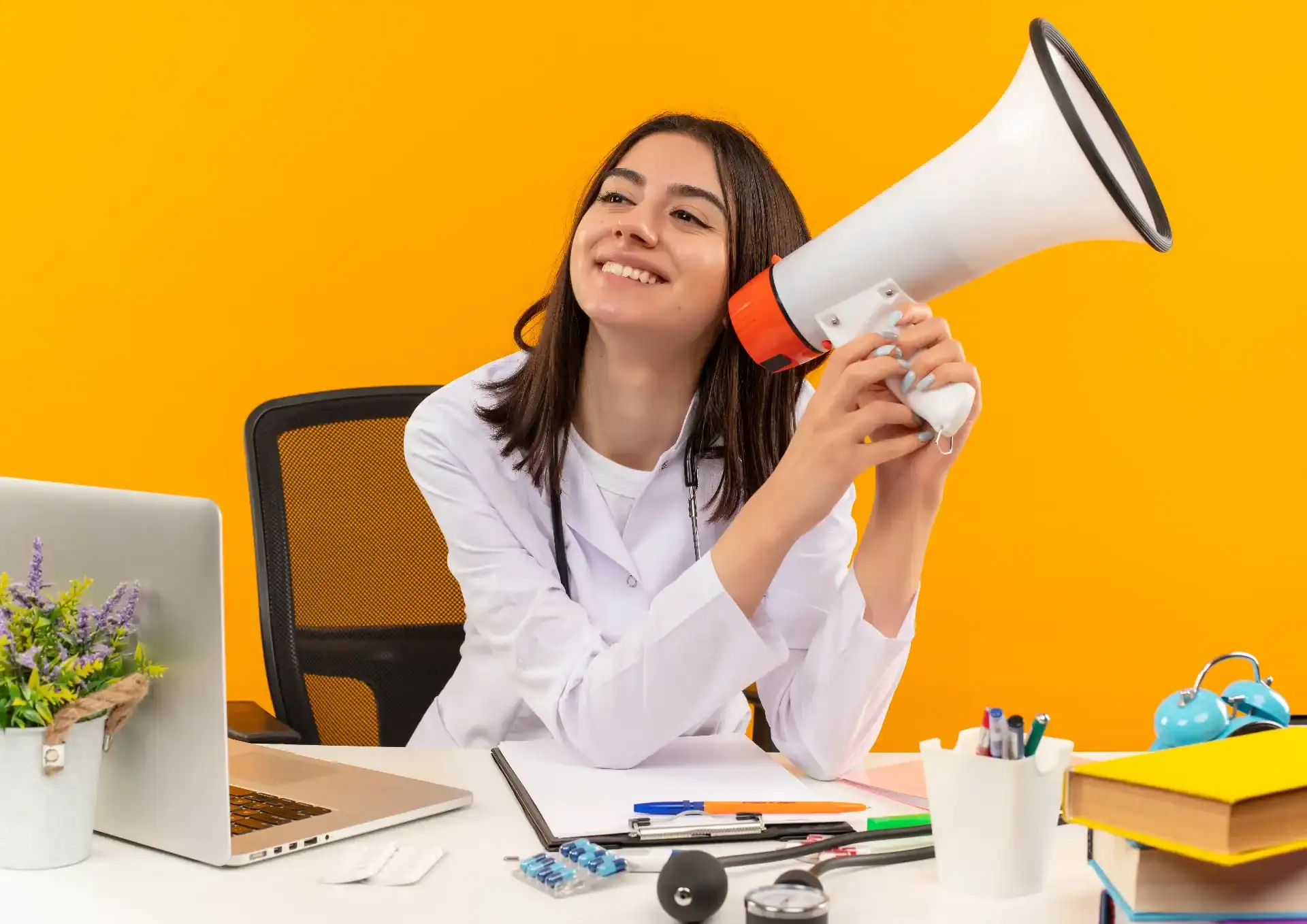 Joven Doctora En Bata Blanca Con Estetoscopio Sosteniendo Megafono Sonriendo Con Cara Feliz Sentado En La Mesa Con Computadora Portatil Y Documentos Sobre Pared Naranja