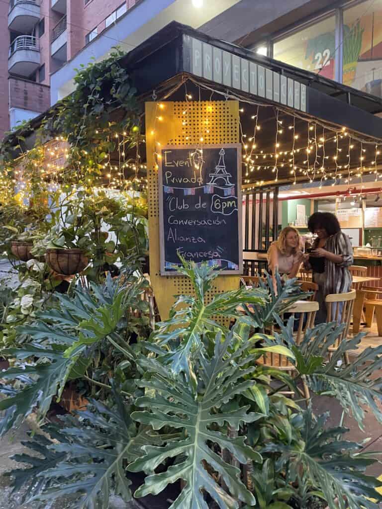 Ambientación de café en exterior con iluminación de guirnaldas, plantas grandes y cartel que invita a eventos privados y clubes de conversación en un ambiente acogedor y vibrante en Bogotá.