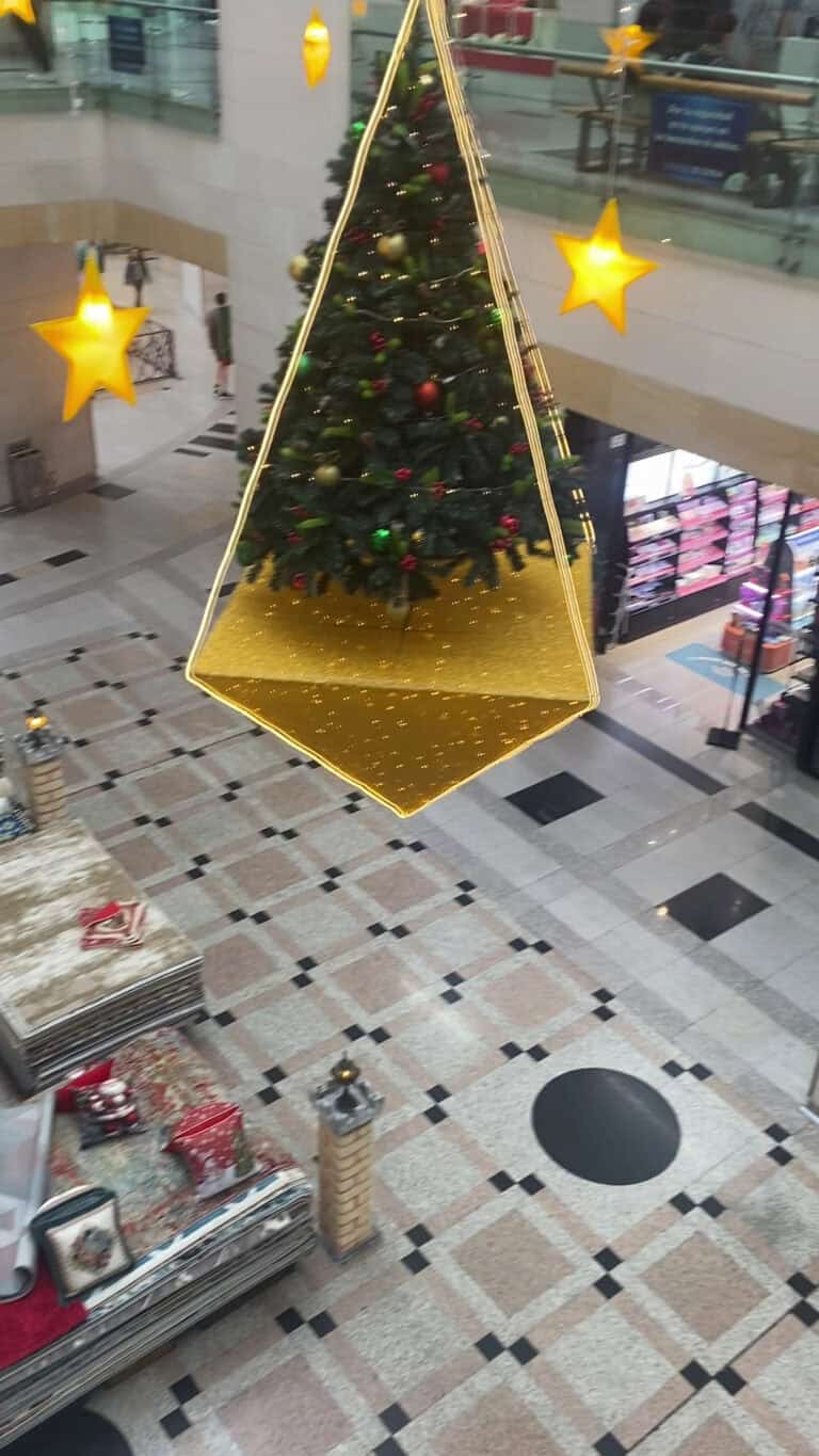 Árbol de Navidad con adornos rojos y dorados, decorado con luces y una base dorada, en un ambiente festivo y comercial.
