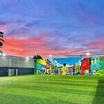 Cancha de fútbol con césped artificial, mural colorido de barrio, cielo al atardecer y malla protectora en urbanización moderna en Colombia.