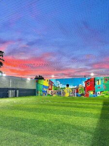 Cancha de fútbol con césped artificial, mural colorido de barrio, cielo al atardecer y malla protectora en urbanización moderna en Colombia.