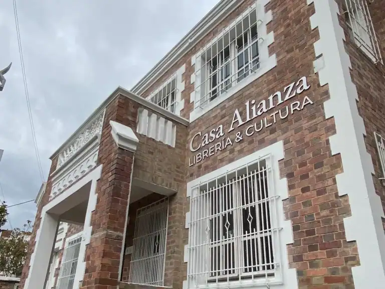 Fachada de la tienda Casa Alianza Librería & Cultura en Colombia, con muro de ladrillos y ventanas con rejas blancas, en un día nublado.