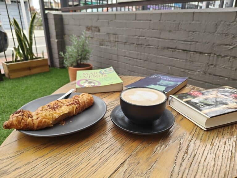 Disfruta de un café con leche acompañado de un croissant en un ambiente relajado, rodeado de libros y plantas en un espacio exterior con vista a la ciudad.