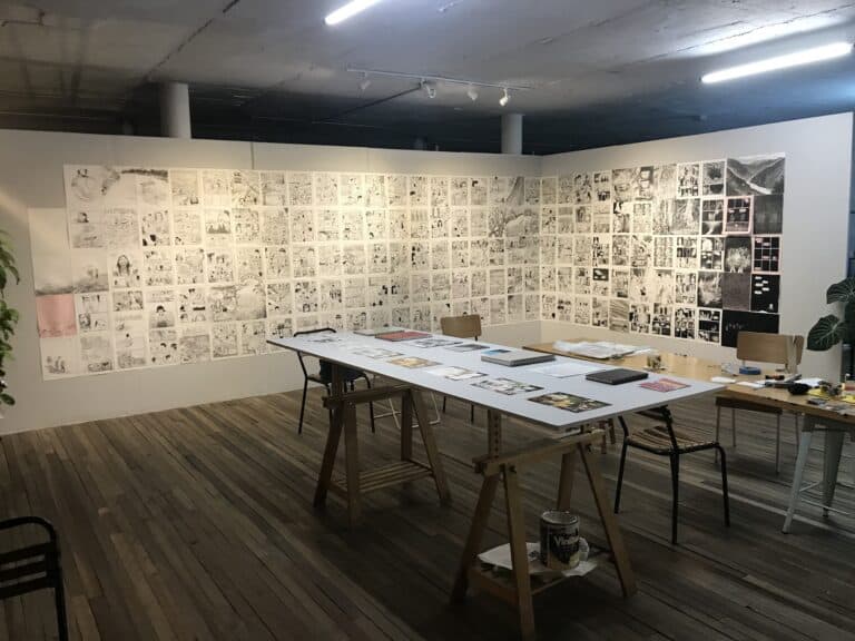 Imagen de una exposición de cómics y dibujos en un espacio de arte en Eventario, con mesas de trabajo y paredes llenas de ilustraciones y obras de arte relacionadas con La Bruja Riso.