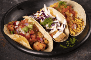Tacos mexicanos con pollo, carne y vegetales en un plato negro, ideales para disfrutar en Braaap Café.