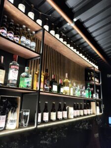 Vista de la barra de bebidas en El Origen Pub, con una variedad de licores y cervezas en estantes de madera y iluminación moderna, ideal para eventos y reuniones sociales.