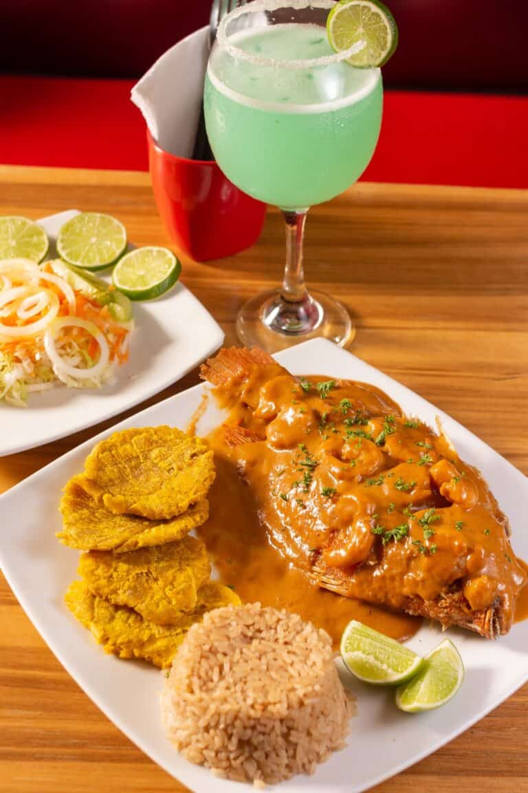 Disfruta de un plato de pescado en salsa con arroz y plátanos fritos en Restaurante El Santuario, acompañado de una bebida refrescante de color verde y limón.
