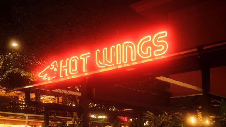 Cartel de neón con el texto "Hot Wings" iluminado en un restaurante, promocionando alitas de pollo picantes en un ambiente nocturno y moderno.