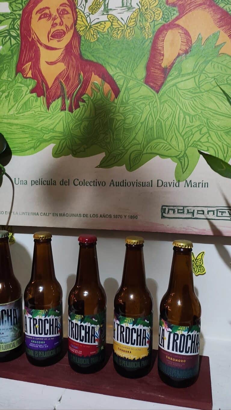 Imagen de botellas de cerveza artesanal La Trocha en un evento cultural en La Casa de la Paz, con un mural colorido de fondo que refleja la cultura y tradición local.