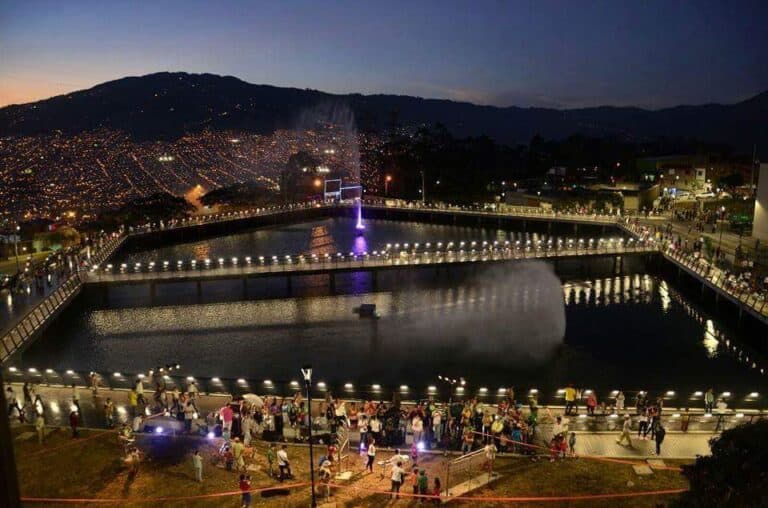 Lago iluminado en la UVA de la Armonía con fuente y visitantes disfrutando del entorno nocturno.