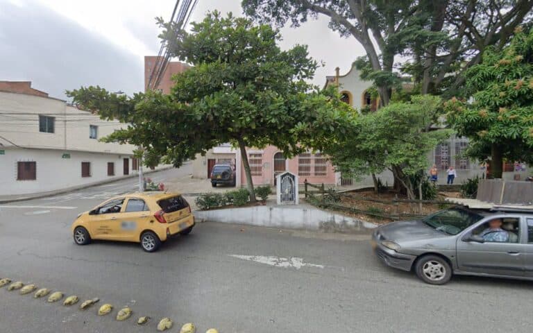 Vista exterior del Café La Musa Rosada con árboles y vehículos en la calle, en un día nublado, en un entorno urbano con edificios y personas en el fondo.