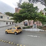 Vista de la fachada de Café La Musa Rosada con árboles y vehículos en la calle, en un día nublado, en un entorno urbano con edificios y personas en el fondo.