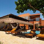 Imagen de una casa de playa con área de descanso, tumbonas y palapa, rodeada de vegetación y con vista al cielo azul, ideal para eventos y vacaciones en la costa.