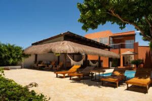 Imagen de una casa de playa con área de descanso, tumbonas y palapa, rodeada de vegetación y con vista al cielo azul, ideal para eventos y vacaciones en la costa.