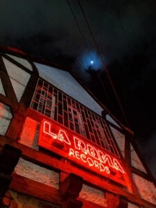 Neón rojo que muestra "La Roma Records" en la fachada de una tienda de discos en la noche, con cielo oscuro y luna visible, creando un ambiente vibrante y nostálgico.