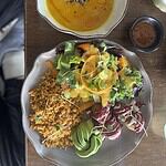 Plato de ensalada con verduras frescas, arroz integral y aderezo, acompañado de una sopa de calabaza y especias, en un ambiente natural y acogedor.