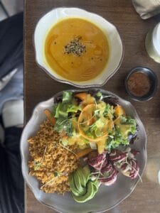 Plato de ensalada con verduras frescas, arroz integral y aderezo, acompañado de una sopa de calabaza y especias, en un ambiente natural y acogedor.